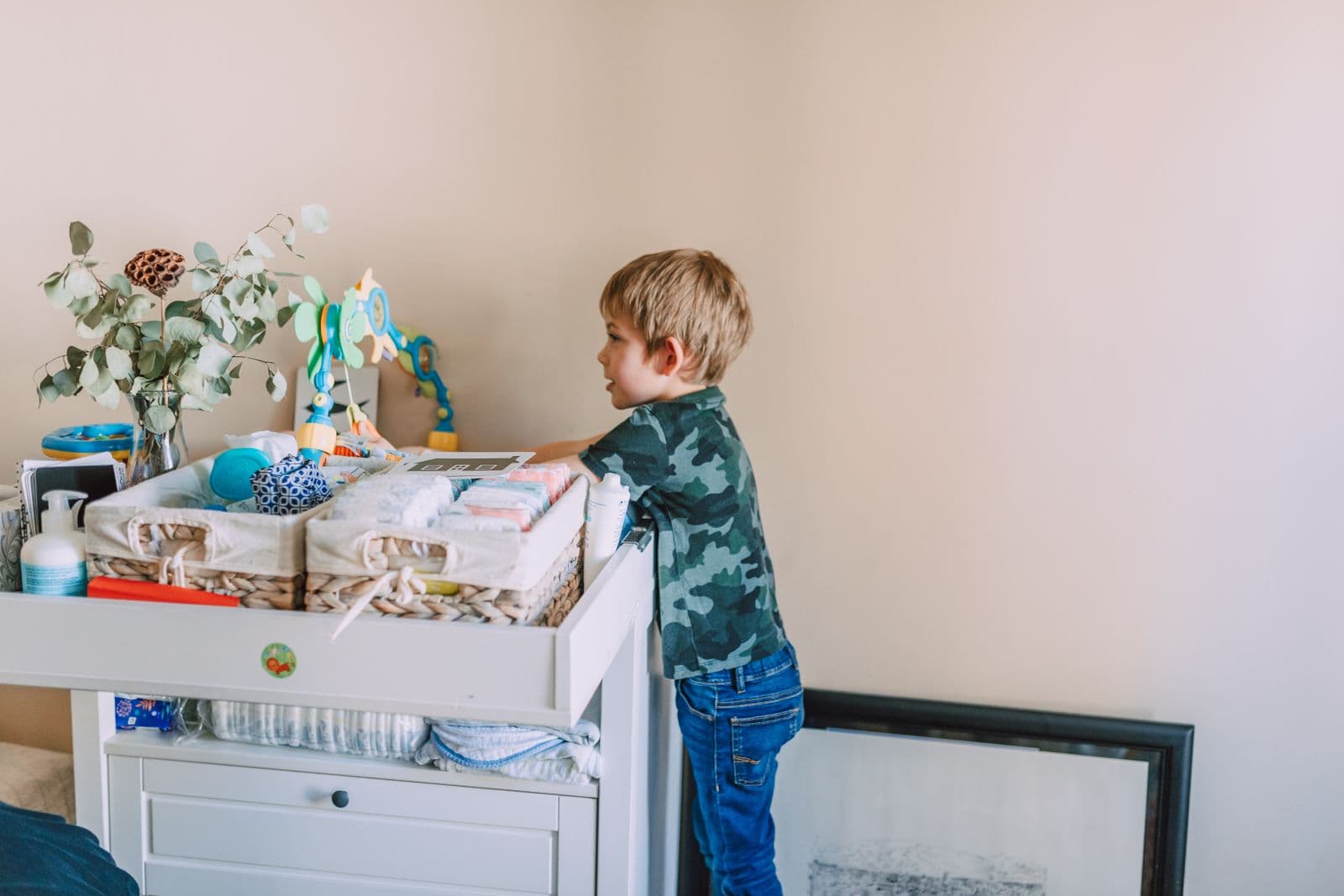 A young child reaching up at a loaded white nursery dresser and changing table against a pale wall