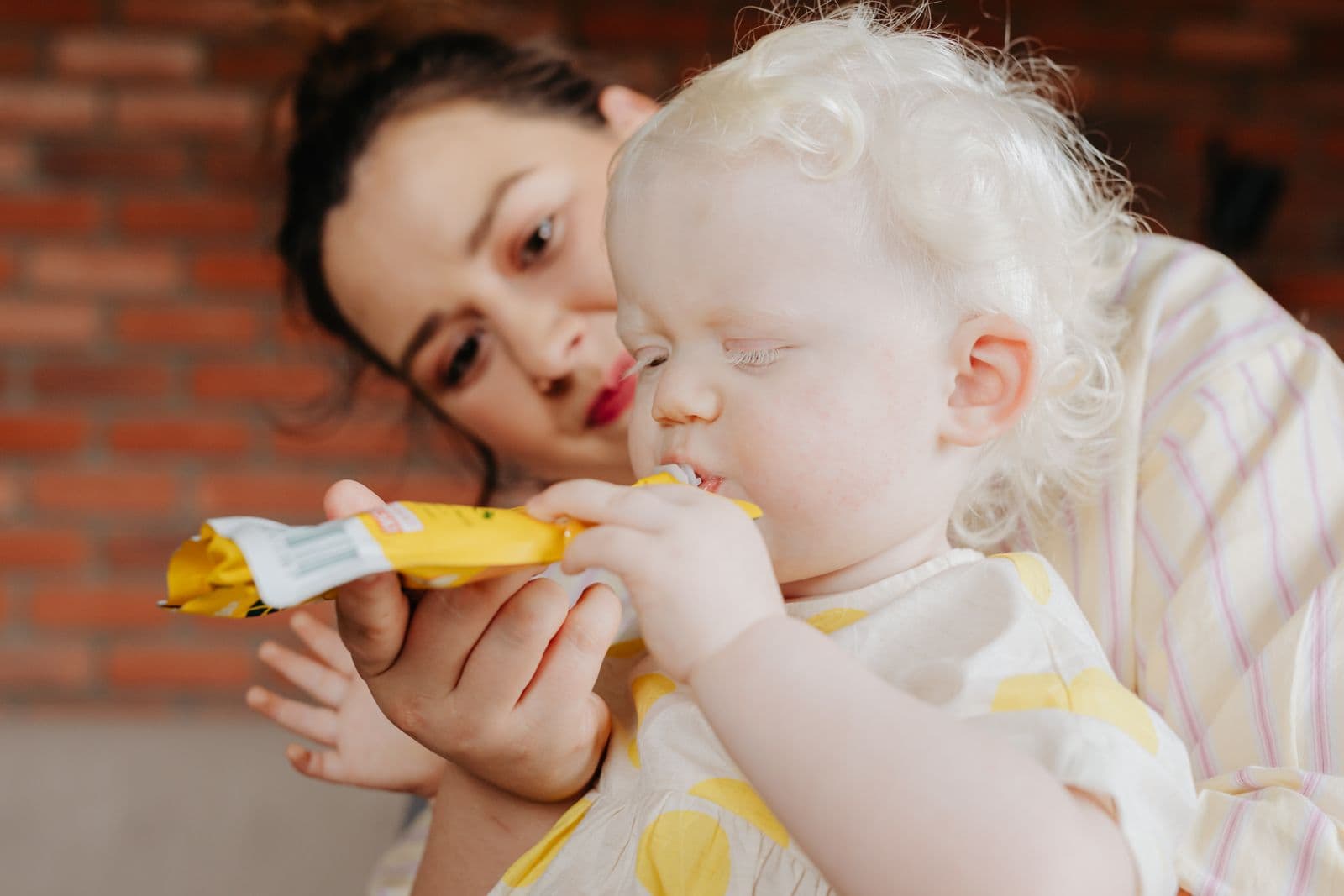 A toddler with pale curly hair drinks from a yellow squeeze pouch while a woman holds the pouch and watches