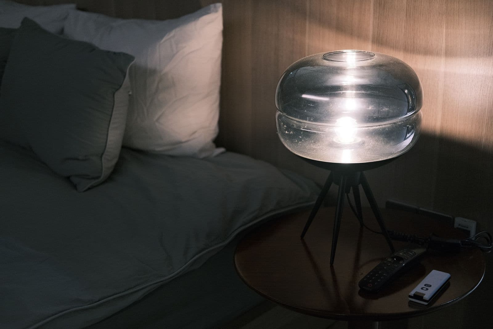 A dim bedside table at night with a glass lamp, a remote and a small white handheld device beside a made bed