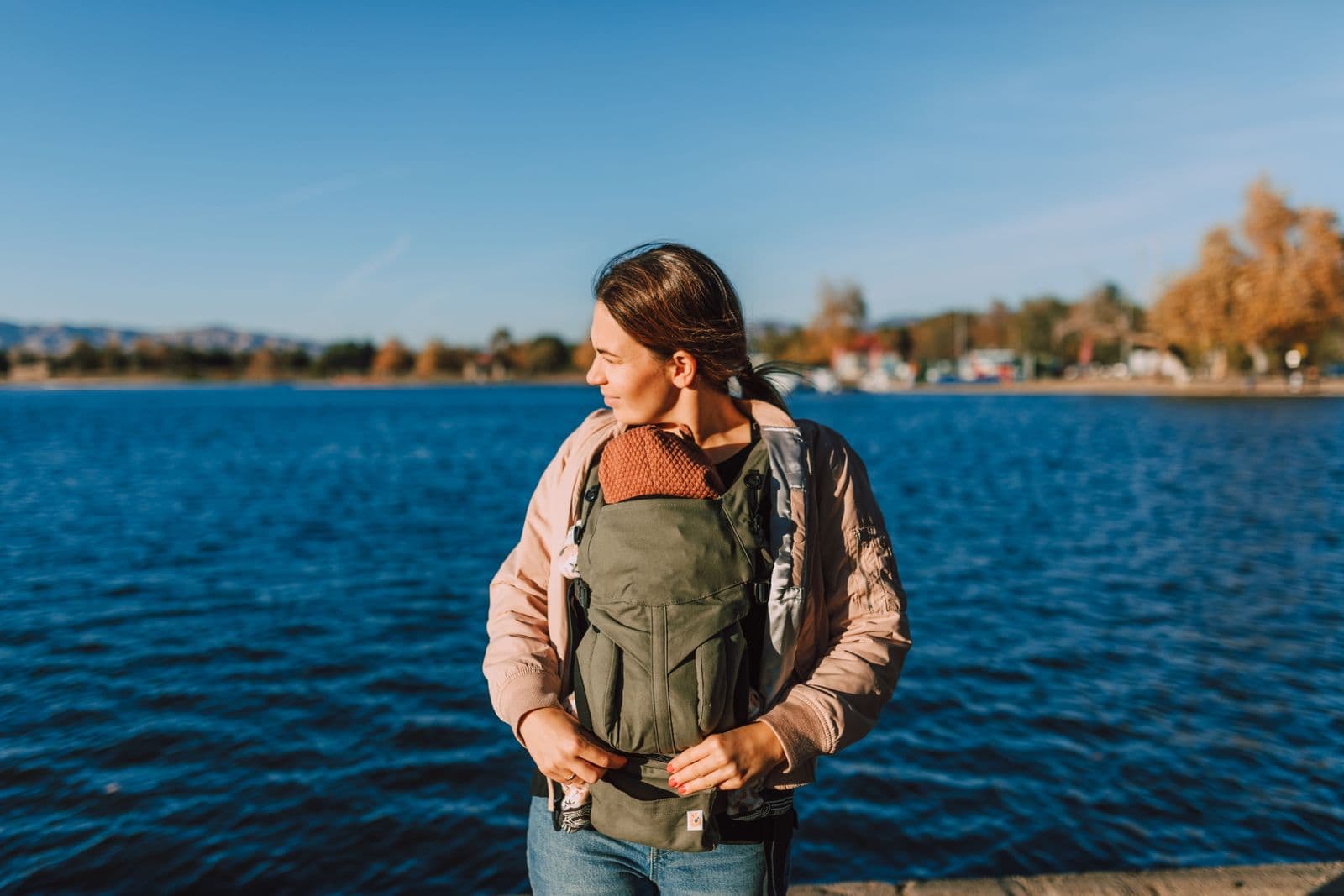 A parent wearing a structured baby carrier outdoors on a bright, sunny day by the water