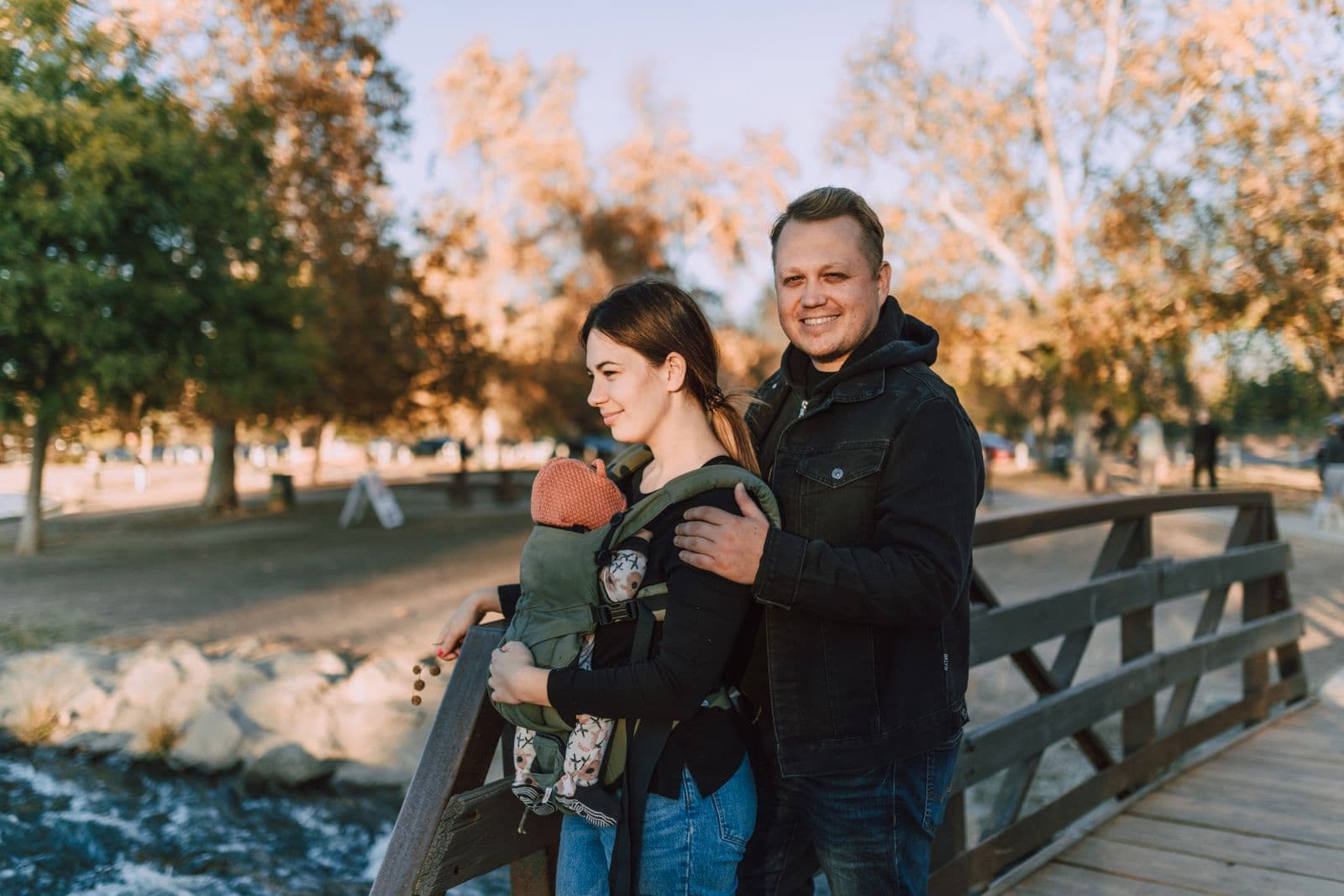 Two parents on a park bridge in autumn with a baby in a structured carrier