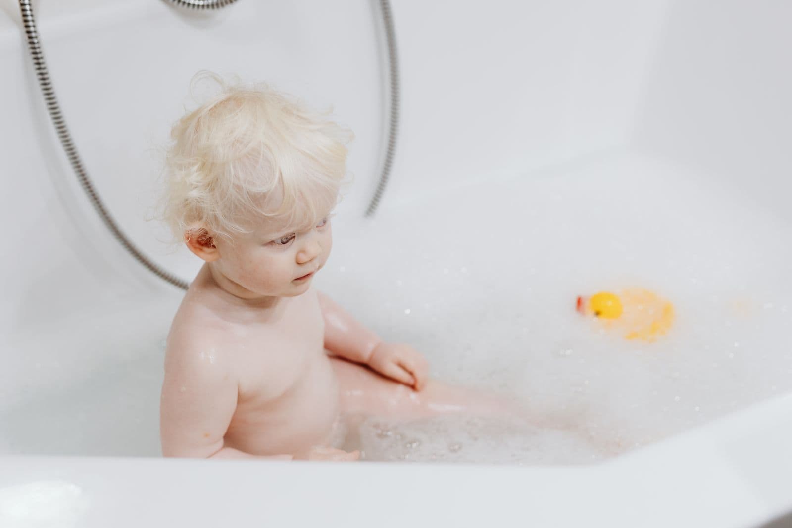 A toddler sitting in a bubble bath in a white tub, looking toward a yellow rubber duck floating nearby