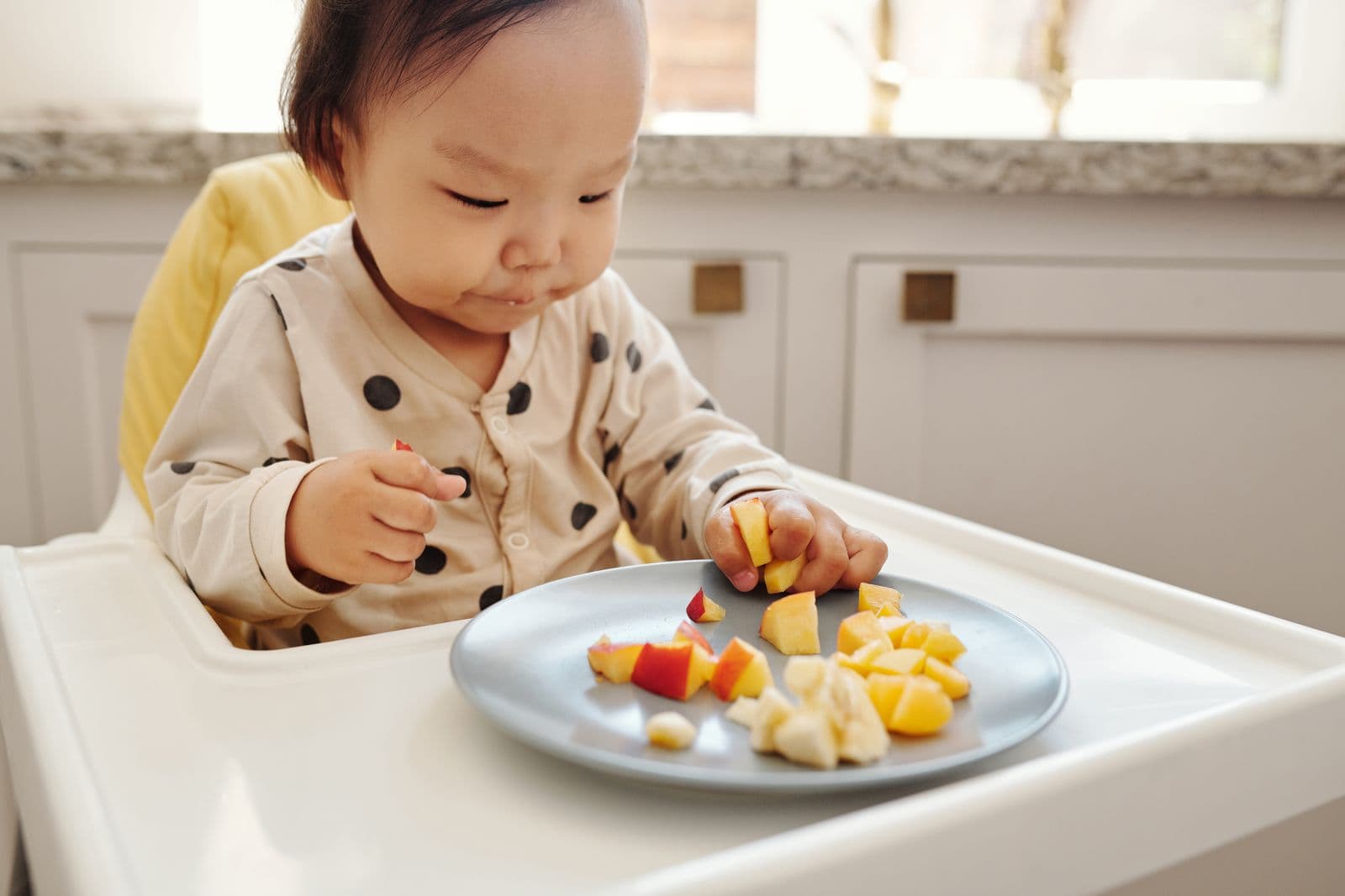 A toddler in a high chair eating cut fruit from a plate