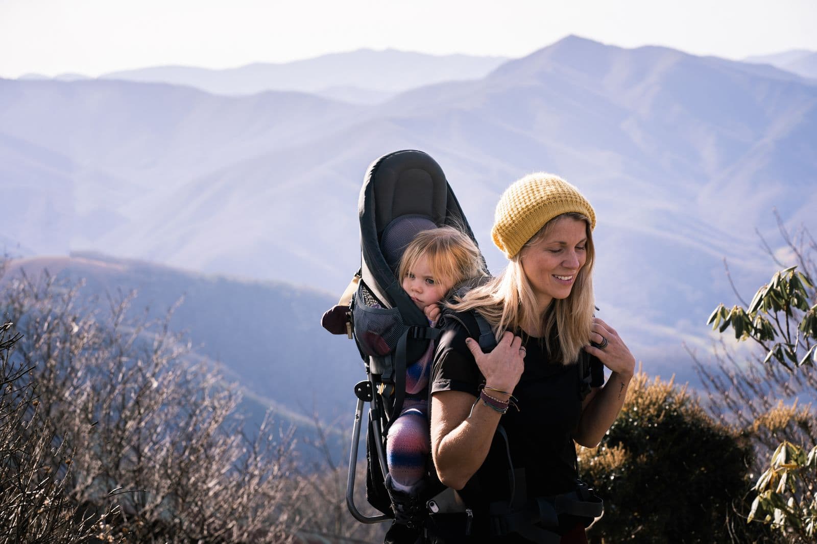A parent on a mountain ridge carrying a toddler in a framed child carrier backpack with a metal kickstand