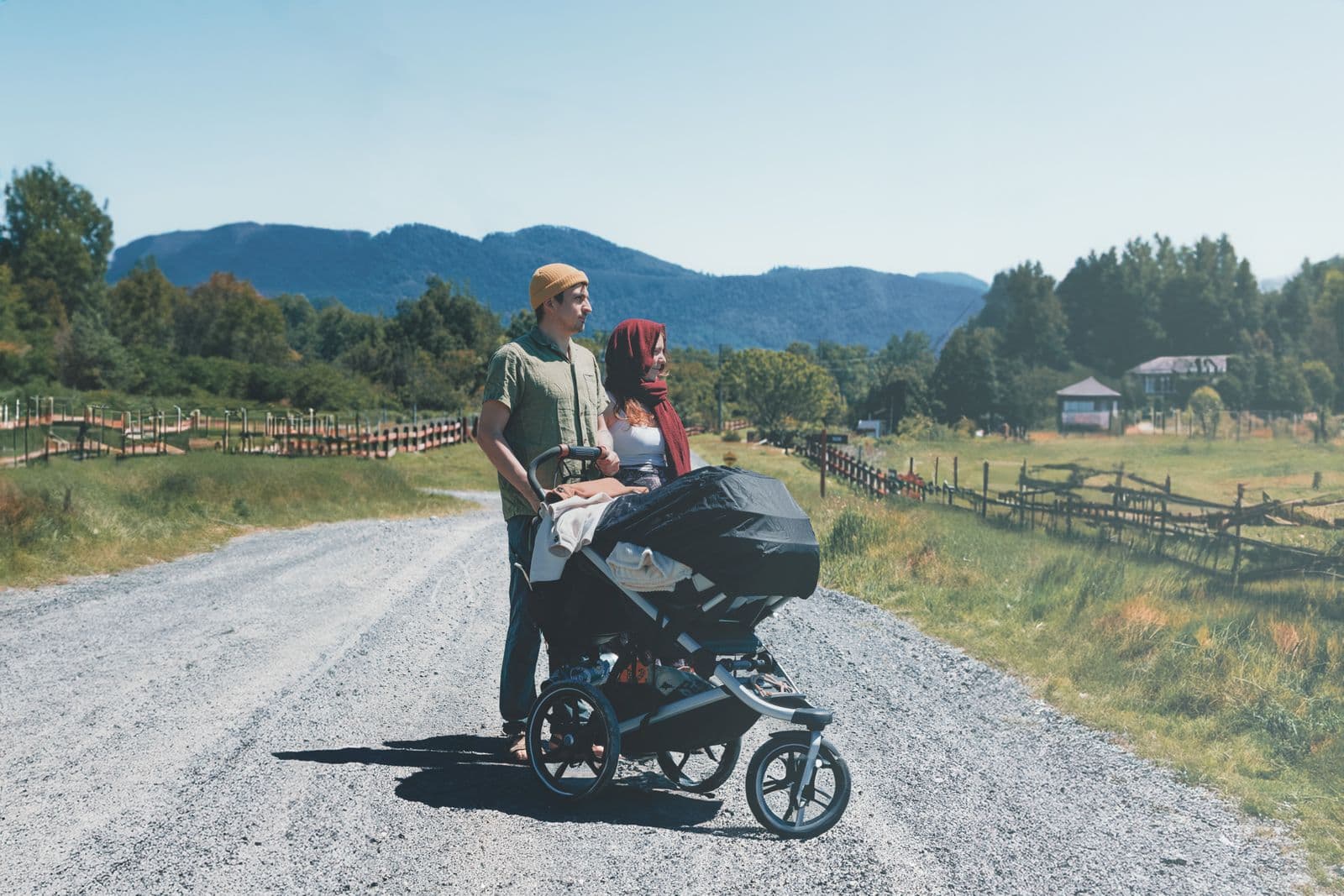 A couple standing on a gravel country road behind a three-wheeled jogging stroller with air-filled tires, mountains in the distance