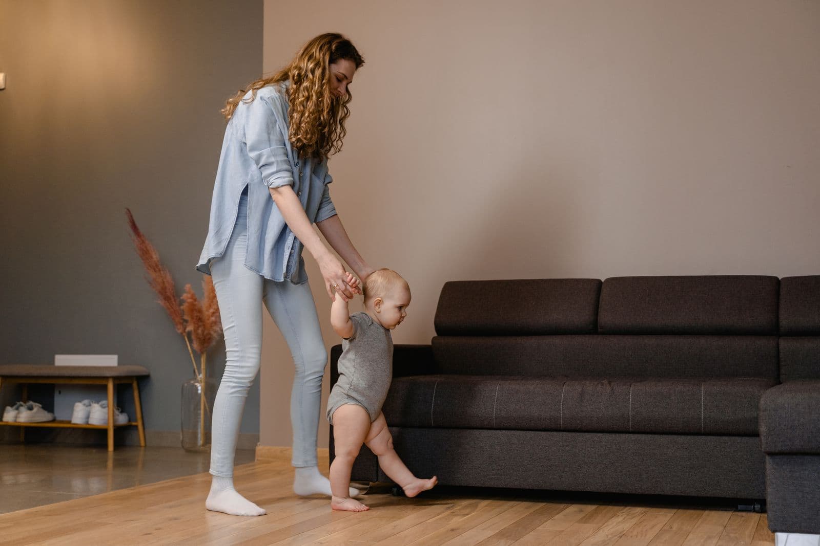 A mother holding a baby's hands as the baby takes first steps across a living room floor