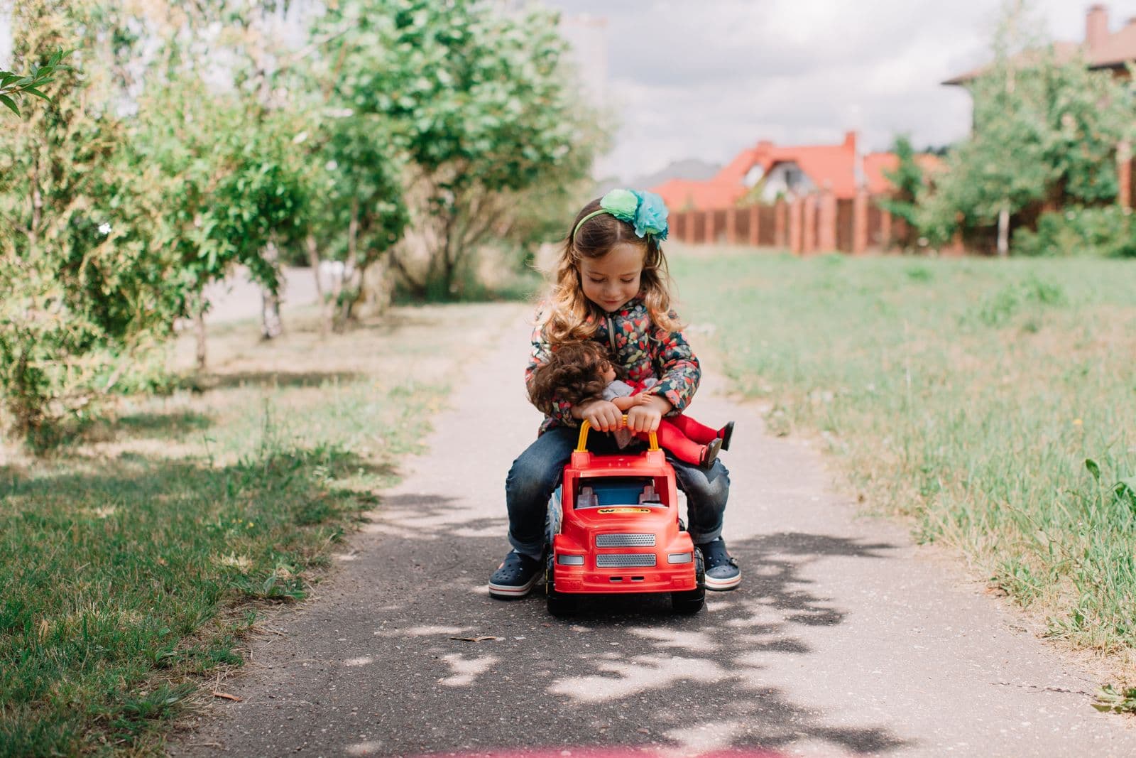 A small girl sits astride a red foot-to-floor ride-on truck on a paved garden path, holding a doll against the handle
