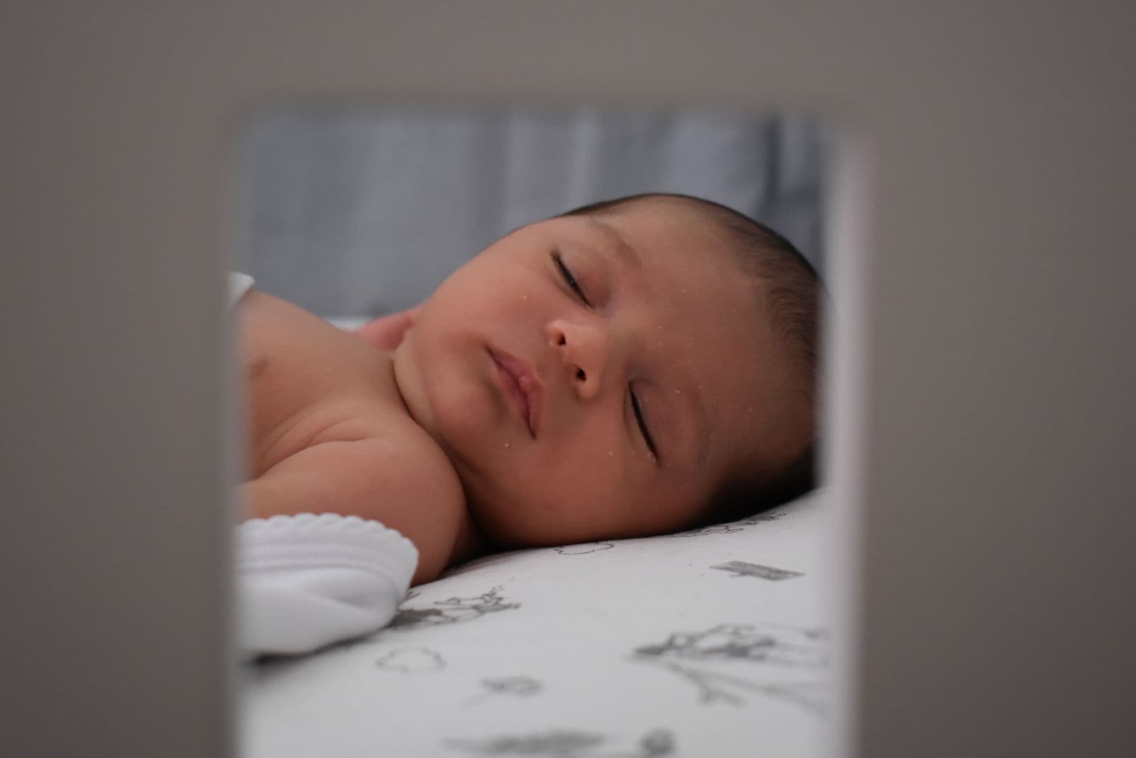 A baby sleeping on their back on a firm, flat mattress, seen through crib rails