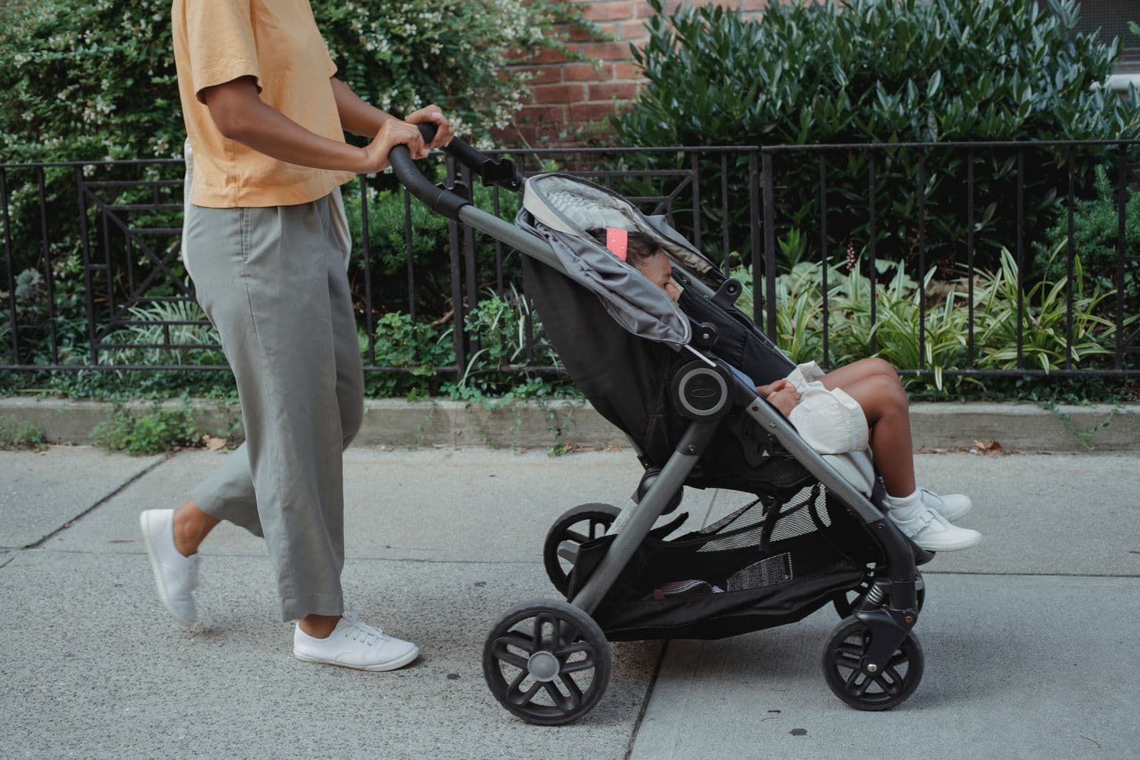 A parent pushing a compact stroller along a city sidewalk with nothing hanging from the handlebar and the basket under the seat