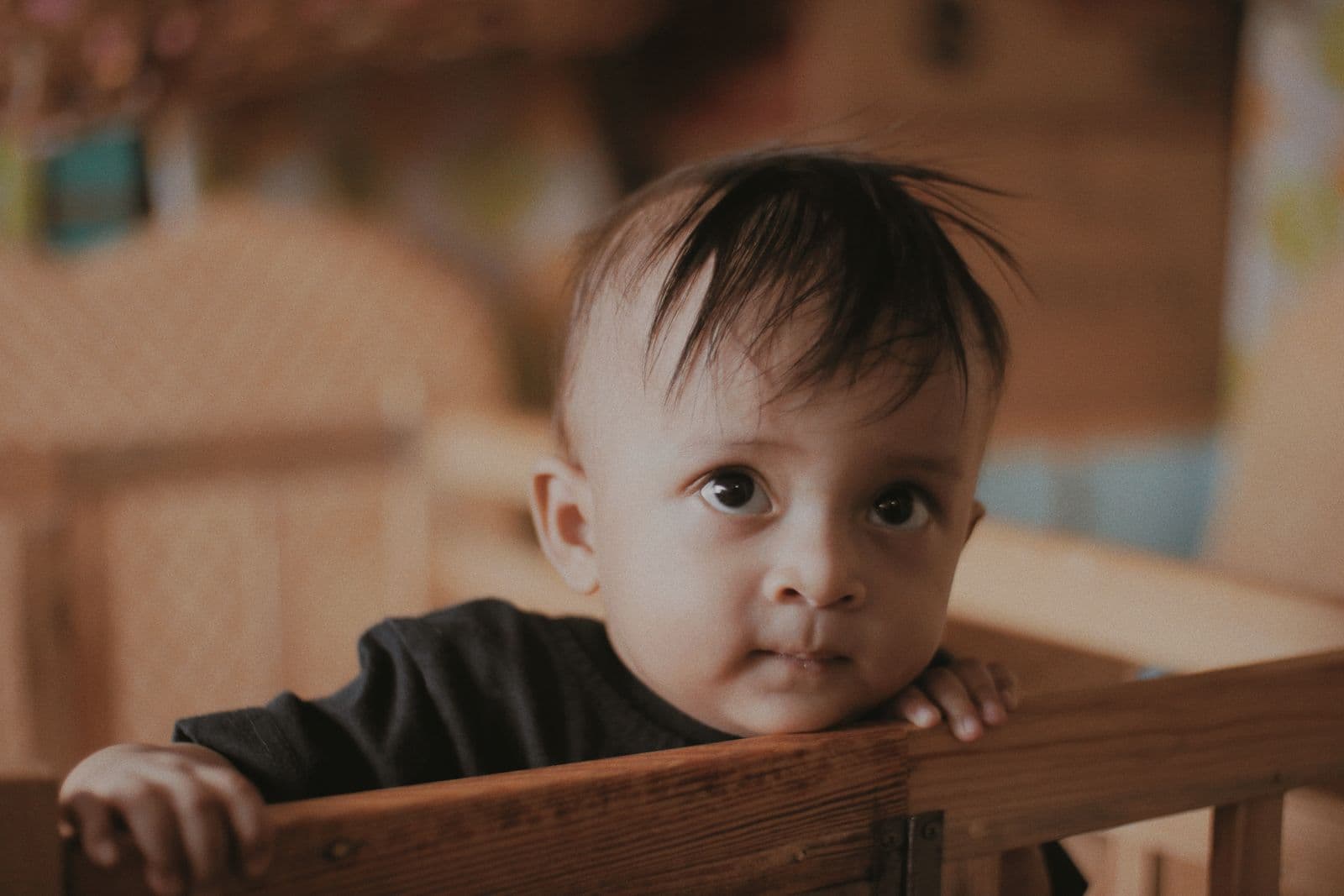 A baby standing at the rail of an older wooden crib, both hands gripping the top rail