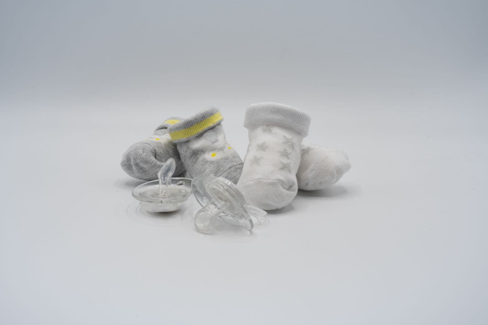 Two clear silicone pacifiers with vented shields lying in front of two pairs of grey and white newborn socks on a white surface