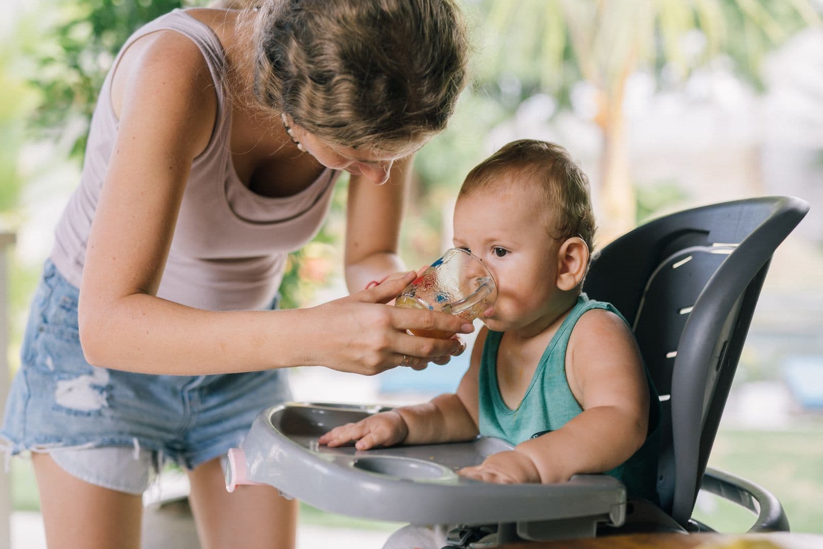 A parent holds a small clear glass cup to a baby's mouth while the baby sits in a grey high chair with a tray