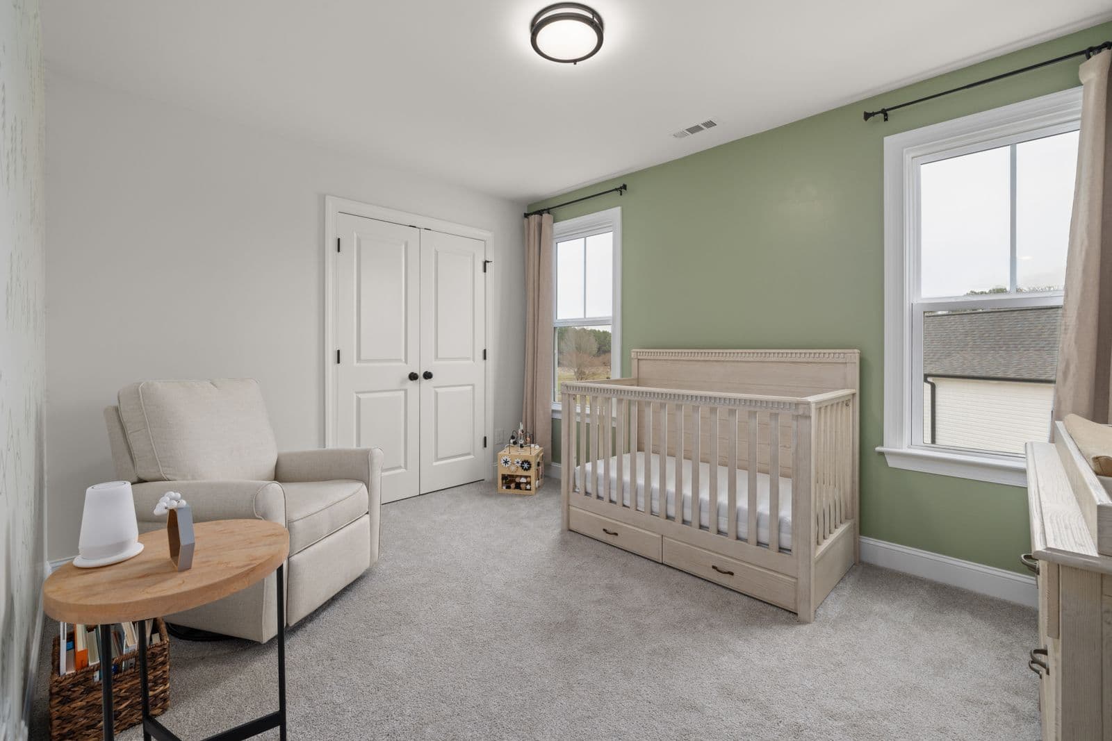 A sage-green nursery with a wooden crib, a glider chair and a small white sound machine on a side table across the room