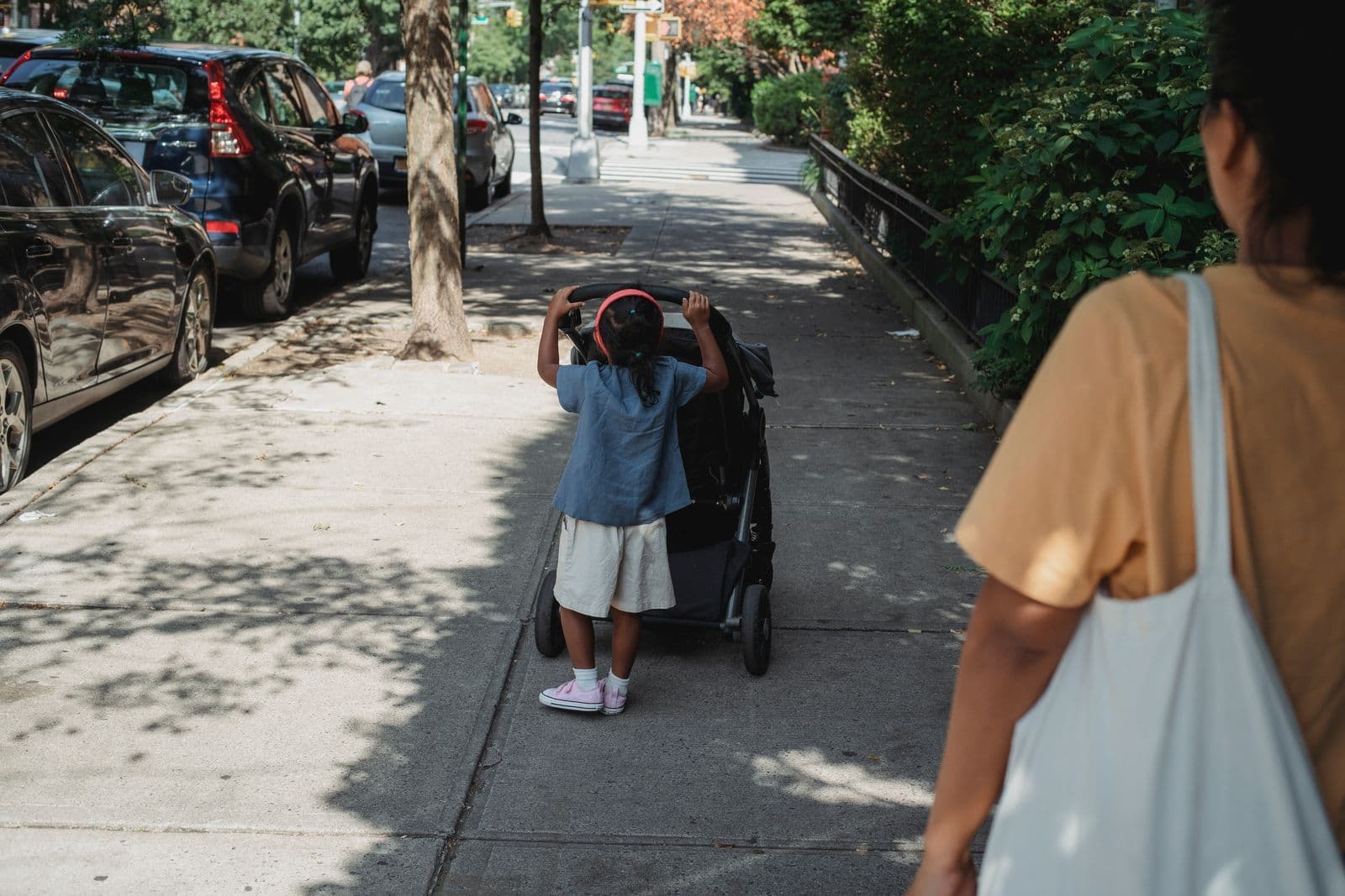 A child pushing a stroller along a sunlit city pavement while an adult walks alongside