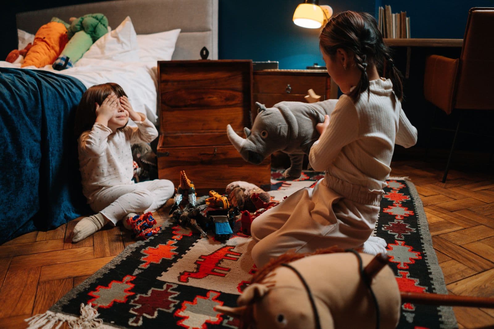 Two young children playing on a rug in a bedroom beside an old wooden storage chest with its lid propped open