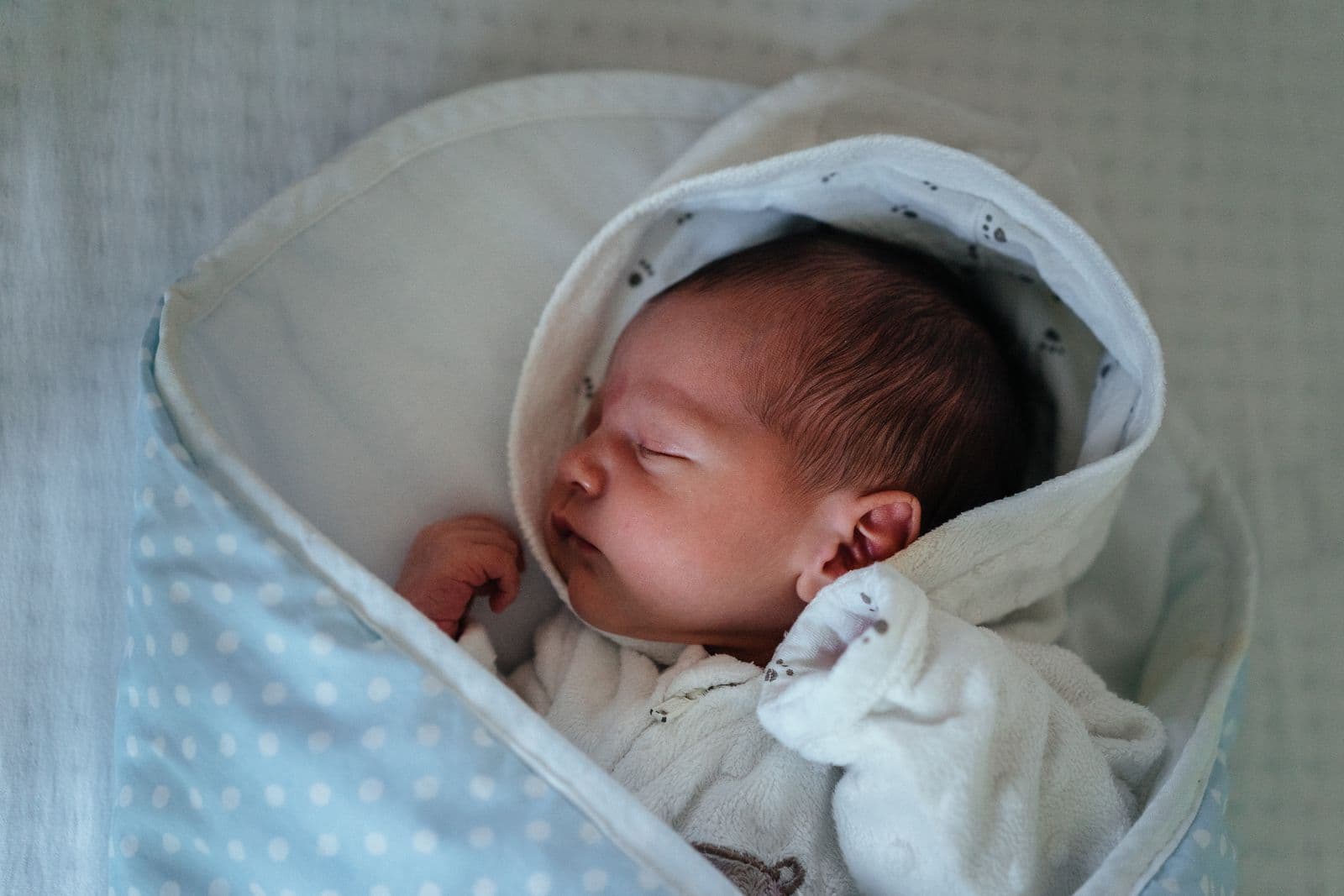A newborn asleep on its back, wrapped in a white and pale blue polka dot blanket with a soft hood, face and one hand uncovered