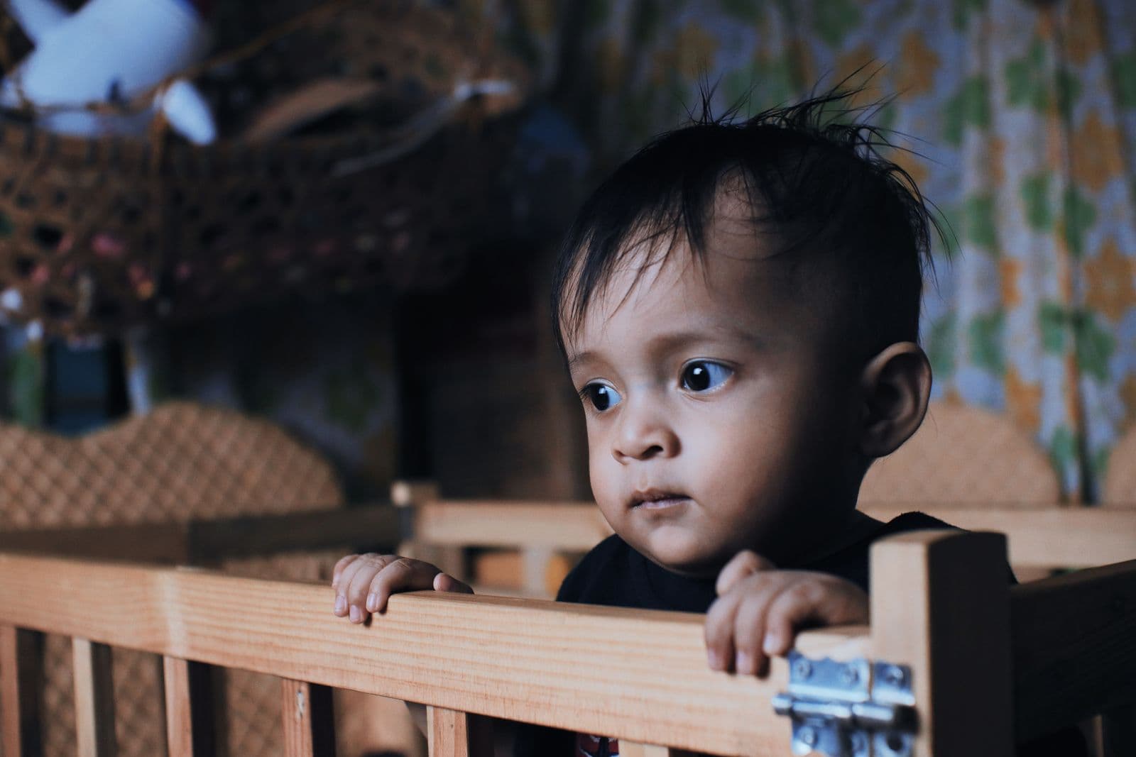 A toddler with dark hair standing in a wooden crib, both hands resting on the top rail, looking out across the room