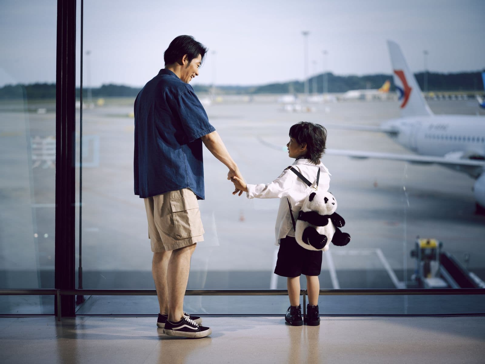 A parent and a small child holding hands at a departure gate window, watching an aircraft on the stand